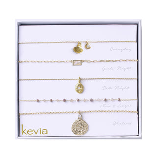 Five-piece gold necklace set featuring shell, crystal, and textured coin pendants with soft gray and pearl beaded accents, packaged in a gift box