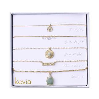 Five-piece gold necklace set featuring textured and gemstone pendants, including a hammered disc, crystal accents, and a green teardrop stone, packaged in a gift box.