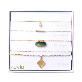 Five-piece gold layered necklace set with a crystal charm, gold bar, green stone accent, and diamond-shaped pendant, packaged in a gift box.