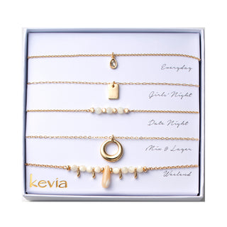 Five-piece gold necklace set with teardrop crystal pendant, gold rectangle charm, mother-of-pearl beads, circular gold pendant, and shell charm, packaged in a gift box.