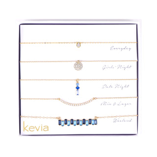 Five-piece gold-tone necklace set featuring sapphire baguettes, teardrop pendant, pavé circle, crystal bar, and sapphire-accented chain.