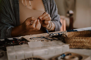 Black woman jewelry designer working at a wooden desk, focused on assembling a necklace with gold chains and natural stone beads in a sunlit studio.