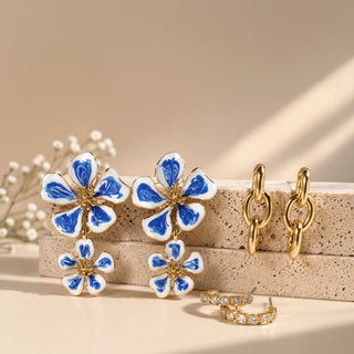 Blue and white enamel flower drop earrings with hand-painted floral petals are displayed beside polished gold link earrings and crystal huggie hoop earrings on stacked travertine stone blocks. 