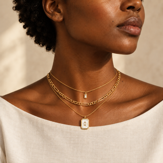 Model wearing three layered gold necklaces with a small rectangular crystal pendant, a link chain and a white rectangular pendant with a crystal center.