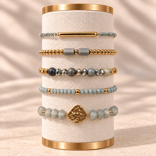 Five grey and gold bracelets with a slim gold bar, grey stone beads, gold beads, and hammered gold centerpiece on a cream display.