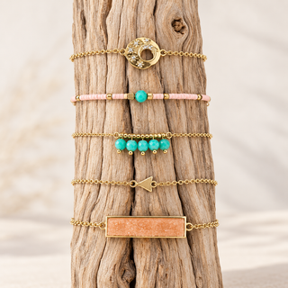 Five gold bracelets with coral rectangular stone, turquoise bead drops, pale pink beads, open textured circle, and arrow charm on wood.