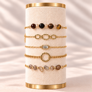 Five gold-toned beaded and chain bracelets displayed on a cream fabric-covered cylindrical stand with gold metal ends, set against a softly draped light beige background.