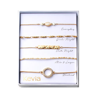 Five piece bracelet set including a chain bracelet with a centered oval cream bead and small white beads, a gold beaded link bracelet, a chain bracelet with a hammered bar centerpiece, a cream heishi bead bracelet with gold accents, and a slider bracelet with an open pavé circle detail, packaged in a gift box.