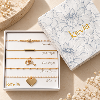 A boxed set of five gold-toned necklaces displayed in an open white gift box next to the matching lid. The set includes Everyday, Date Night, Mine & Luger, Girls Night, and Weekend bracelets, each with a different design.