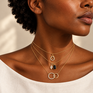 A close-up of a model wearing a layered gold necklace set with three distinct pendants: a crescent moon and star, a teardrop with a black stone, and two interlocking rings with clear stones.
