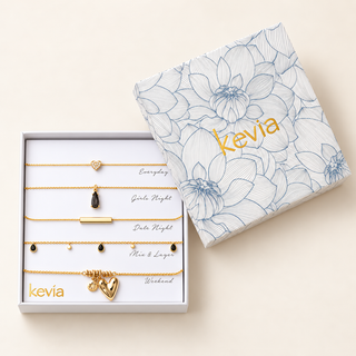 A boxed jewelry gift set labeled kevia, shown with the white presentation box open and the matching floral gift box beside it on a warm cream background. The set contains five delicate gold-toned necklaces with different pendant/charm designs and handwritten color/style labels: Everyday, Girls Night, Date Night, Mine & Yours, and Weekend.