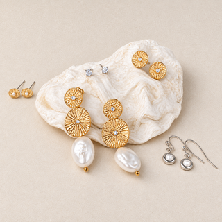 Five pairs of mixed metal earrings with gold sunburst pearl drops, gold textured studs, clear studs, and silver pearl drops on a shell and cream surface.