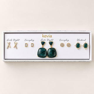 Five-pair gold earring set with deep green stone drops and clusters, clear rectangular studs, crossed bars, and pavé discs in an open Kevia box.