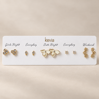 Five-pair gold earring set on a Kevia card with white enamel flowers, teardrop and square studs, crystal clusters, and floral climbers.