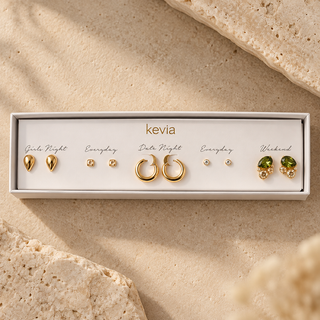 Five-pair gold earring set with green crystal clusters, gold teardrop studs, champagne square studs, clear round studs, and hoops in an open Kevia box.