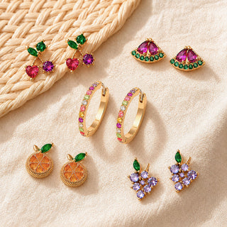 Five pairs of colorful earrings, including cherry, watermelon, rainbow hoop, orange slice, and grape designs, are arranged on textured ivory fabric beside a woven straw mat with polished gold settings and vibrant gemstones.