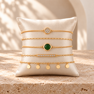 Five gold bracelets with a green round stone, pavé circle, crystal bar, fine chain, and disc drops on a cream cushion.