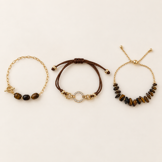 Three delicate bracelets arranged side by side on a light cream background: a gold chain bracelet with three brown tiger's eye beads and a toggle clasp, a brown cord bracelet with gold hardware and a small clear stone circle, and a gold chain bracelet with multiple brown tiger's eye beads.