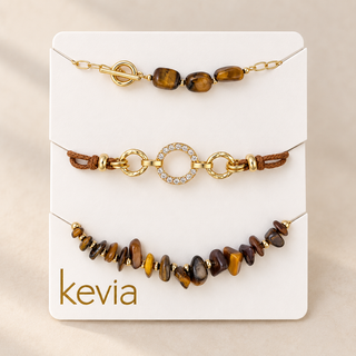 Three delicate gold-toned bracelets displayed on a white card labeled 'kevia'. The top bracelet features polished tiger's eye beads and a toggle clasp.