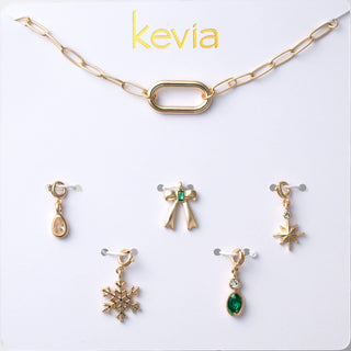 Six-piece gold holiday charm necklace set with paperclip chain, snowflake, bow, teardrop, starburst, and green stone charms, packaged in a gift box.