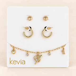 A gold jewelry set on a white display card, including two stud earrings, two hoop earrings, and a delicate chain necklace with small charms, one shaped like a baseball and one with a '1' charm. The brand name 'kevia' is printed in gold on the card.