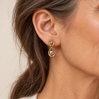 A close-up of a woman's ear wearing a gold drop earring with two oval smoky gemstones.