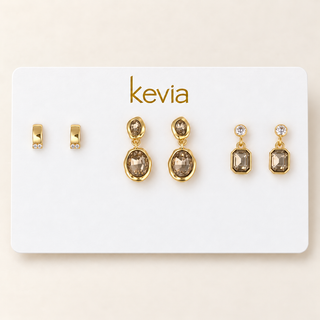 A set of gold-toned earrings displayed on a white Kevia brand card against a light beige background. The set includes small gold huggie hoop earrings with clear stones, a pair of gold drop earrings with oval smoky brown stones, and a pair of gold drop earrings with rectangular smoky brown stones and a clear top stone.