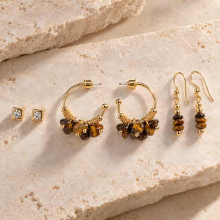 three pairs of gold earrings with cubic zirconia and tiger's eye beads. one pair of posts, a pair of oops and a dangle pair laying on a beige stone background.