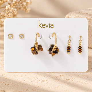 A jewelry display card from Kevia featuring three pairs of gold-toned earrings, including two pairs with brown tiger's eye beads, two small square stud earrings with clear stones, and two beaded drop earrings, set against a warm sandy background.