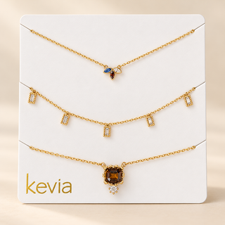 Three gold-tone jewelry pieces displayed on a white branded card labeled kevia. The set includes a delicate chain necklace with a small blue and brown pendant, a charm-style chain with clear rectangular stones, and a longer chain with a larger brown gemstone pendant and small clear floral accents.