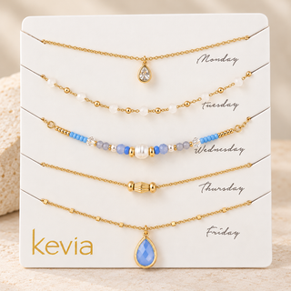 Five gold-toned beaded necklaces displayed on a white card labeled Monday through Friday, with the brand name kevia at the bottom left. The necklaces include pearl and blue bead accents, and two have teardrop-shaped blue and clear stones.
