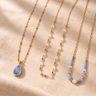 Three delicate gold-tone necklaces arranged diagonally on a textured beige fabric background. The left necklace features a teardrop-shaped light blue stone pendant.