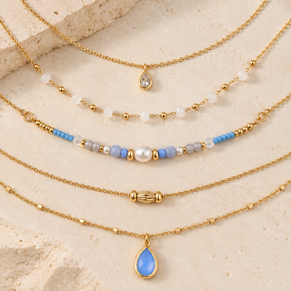 Five layered gold-tone necklaces with various bead and pendant designs, including teardrop pendants and beaded strands with light blue, white, and pearl accents, arranged on a textured beige stone background.
