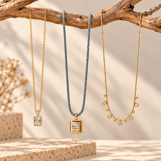 Three delicate necklaces displayed on a rustic wooden branch: two gold-tone chains with square crystal pendants and one blue braided cord necklace with a gold square pendant, set against a warm beige background with soft shadows.