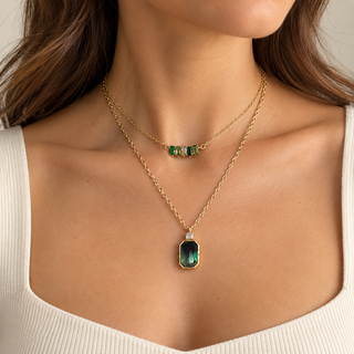 A close-up of a model wearing a double-layered gold necklace set. The shorter chain features a small bar with three emerald-cut green stones, while the longer chain holds a larger emerald-cut green pendant with a gold bezel and a small clear accent stone.