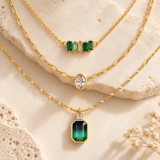 Three gold necklaces arranged on a light beige stone surface: a top necklace with four green emerald-cut stones, a middle necklace with one oval clear stone, and a bottom necklace with a larger green emerald-cut pendant and a small clear stone.