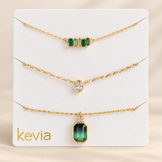 Three gold chain necklaces displayed on a white Kevia brand card. The top necklace has four green emerald-cut gemstones, the middle has a single oval clear gemstone, and the bottom has a larger rectangular green emerald-cut pendant with a small clear accent stone.