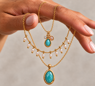 A close-up of a hand holding three layered gold necklaces. The necklaces feature a teardrop-shaped turquoise pendant, a larger oval turquoise pendant with a faceted stone, and delicate strands of white faux pearls with small gold beads.
