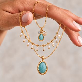 Hand holding three gold necklaces with an aqua teardrop and crystal charm cluster, dangling white pearls and a large oval aqua pendant.