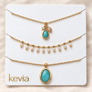 Three gold-tone layered necklaces displayed on a white card labeled 'kevia'. The necklaces include a small charm with a turquoise stone, a strand of white faux pearls with gold beads, and a larger oval turquoise stone pendant.