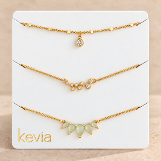 Three gold-tone delicate necklaces displayed on a white Kevia brand card, featuring a teardrop crystal pendant, a crystal cluster, and a row of pale green teardrop gemstones.