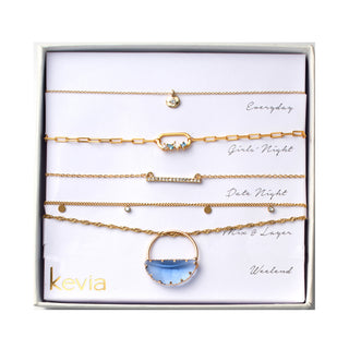 Five piece necklace set packaged in a gift box featuring a gold necklace with a small round charm pendant, a gold paperclip chain necklace with small blue crystal accents, a slim gold bar necklace with clear crystal detailing, a gold chain necklace with evenly spaced small round disc charms, and a gold necklace with a circular light blue stone pendant framed in gold.