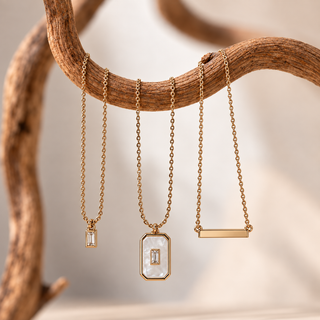 Three gold-tone necklaces with rectangular pendants are displayed hanging from a curved wooden branch. The left two pendants feature clear gemstones, and the center pendant is a white mother-of-pearl rectangle with a matching clear stone in the middle.