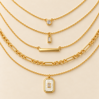 Five layered gold necklaces with various pendant designs, including a bezel-set clear stone, an emerald-cut stone, a bar, and a rectangular pendant with a white inlay, arranged on a light beige background.