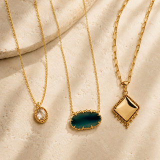 Three gold necklaces with a clear oval charm, green stone pendant, and diamond-shaped gold pendant on cream stone.