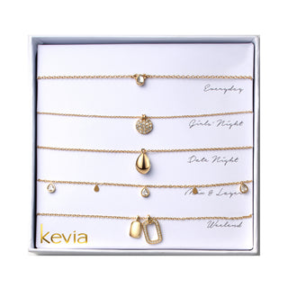 Five piece necklace set packaged in a gift box featuring a gold necklace with a small polished knot charm, a gold necklace with a round pavé crystal disc pendant, a gold necklace with a smooth teardrop shaped pendant, a gold chain necklace with small clear crystal drop charms, and a gold necklace with two charms including a rectangular pavé crystal frame and a smooth gold tag.