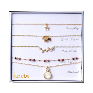 Five piece necklace set packaged in a gift box featuring a gold necklace with a small crystal flower charm, a gold necklace with a mixed charm cluster including a white pearl bead, a faceted amber brown stone, and a clear crystal accent, a gold necklace with a curved floral crystal motif, a gold chain necklace with alternating faceted garnet red and clear beads, and a gold necklace with a round clear crystal pendant framed in gold.