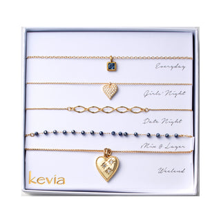 Five piece necklace set packaged in a gift box featuring a gold necklace with a square sapphire blue crystal pendant, a gold necklace with a crystal pavé heart charm, a gold open link chain necklace, a gold chain necklace with sapphire blue faceted bead accents, and a gold heart locket necklace decorated with small clear crystal details.