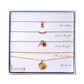 Five piece necklace set packaged in a gift box featuring a gold necklace with a slim vertical ruby red crystal pendant, a gold curved crystal bar necklace, a gold necklace with a square ruby red charm and small crystal key accent, a fine gold chain with evenly spaced spacer details, and a gold textured heart medallion necklace with a centered clear crystal.
