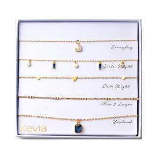 Five piece necklace set packaged in a gift box featuring a gold crystal accented heart charm necklace, a gold station necklace with alternating sapphire blue baguette charms and round clear crystal drops, a gold chain with three small polished heart stations, a slim gold chain with evenly spaced rectangular spacer accents, and a gold rectangular sapphire blue pendant necklace.
